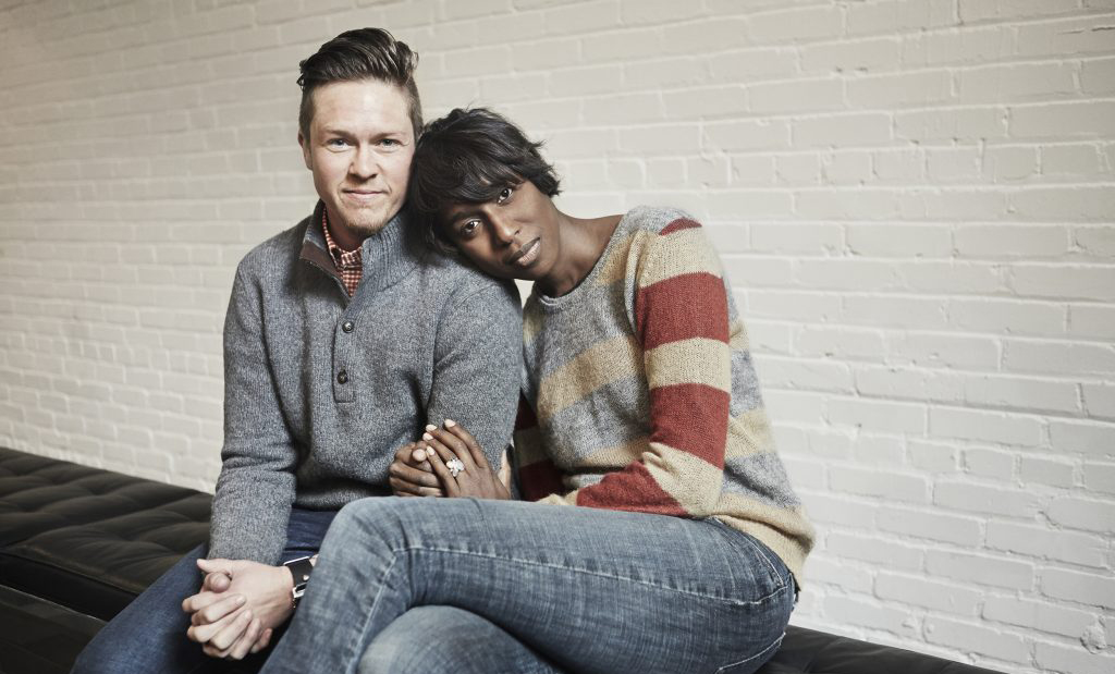White trans-masculine person and Black trans-feminine person sitting together, looking into camera.
