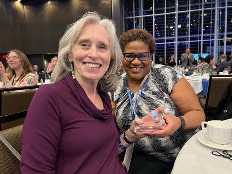 Photo of Kate Callery sitting at a dining table at the Ray Denison Award ceremony. She is a white woman with grey hair. Next to her is Angie Hale, a Black woman with short hair. They look like they're having fun. :)
